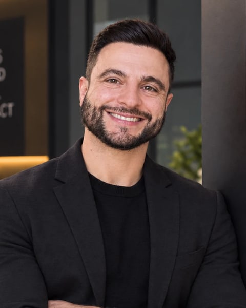 Luiz Hogrefe, smiling, arms crossed, in a dark blazer over a black knit top, leaning against a dark wall